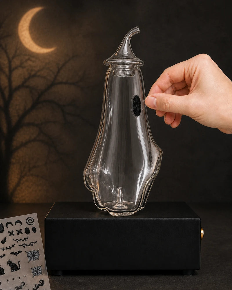 A hand applying one included Halloween eye sticker to the blank ghost-shaped glass bottle on the black MINI base